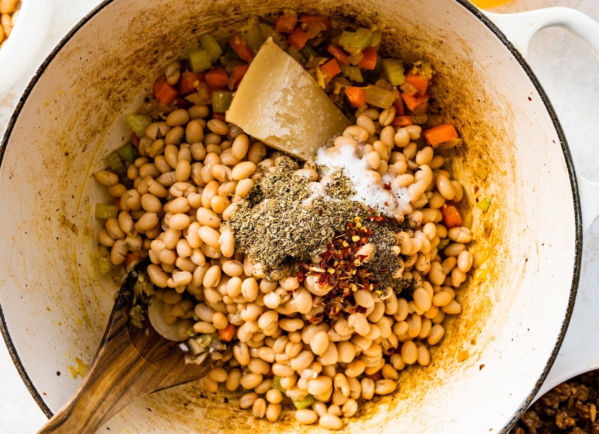 white beans, herbs, spices, and parmesan rind being added to white soup pot with wooden spoon.