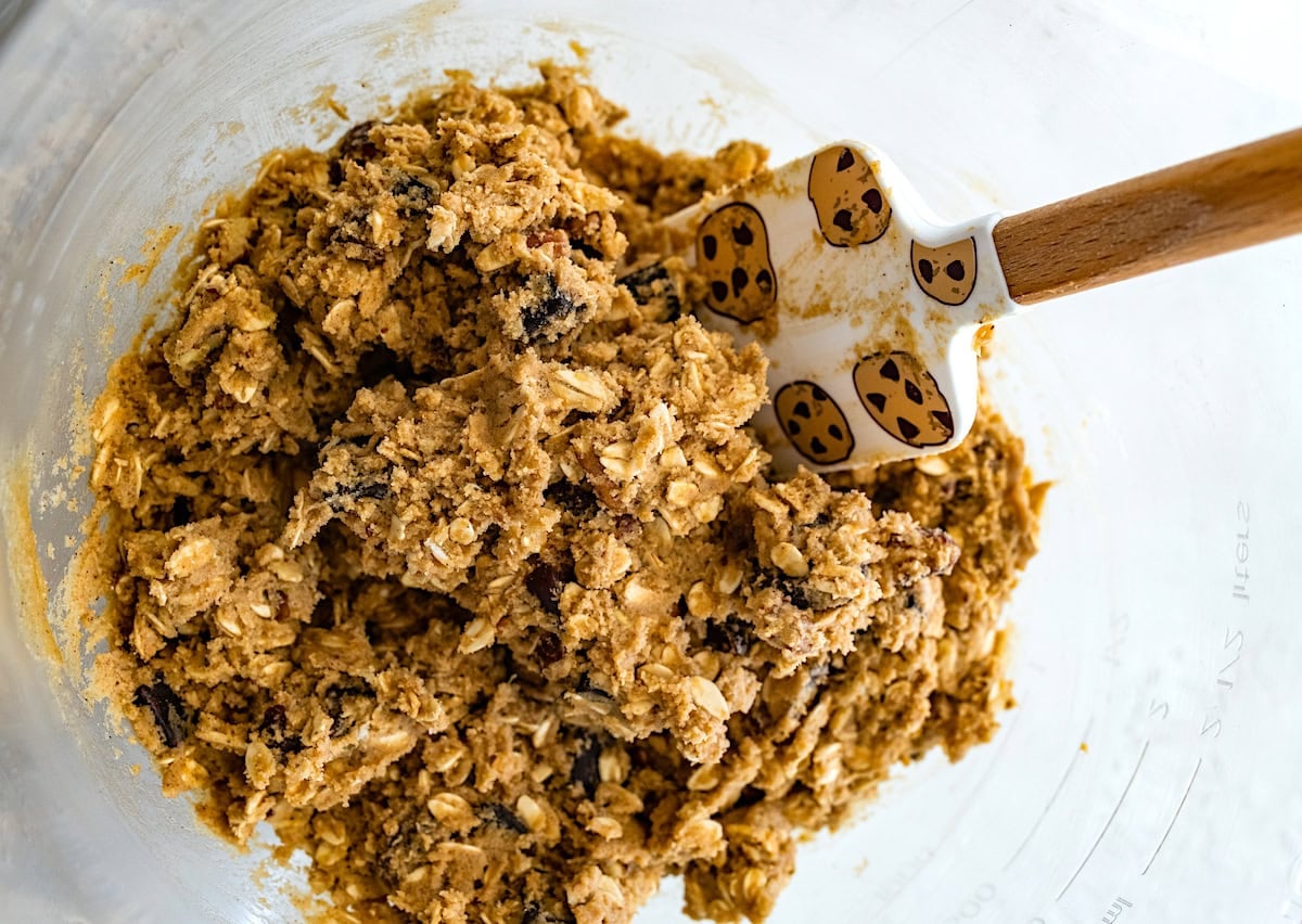 brown butter oatmeal cookie dough with pecans, dates, and chocolate chunks in mixing bowl with spatula.