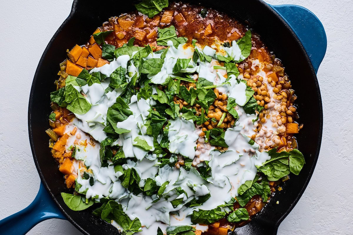 spinach and coconut milk being added to sweet potatoes and lentils in cast iron skillet.