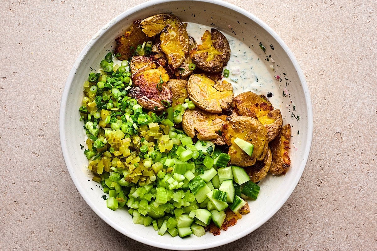 smashed potatoes, diced cucumber, diced celery, pickles, green onion, and creamy herb dressing in bowl.