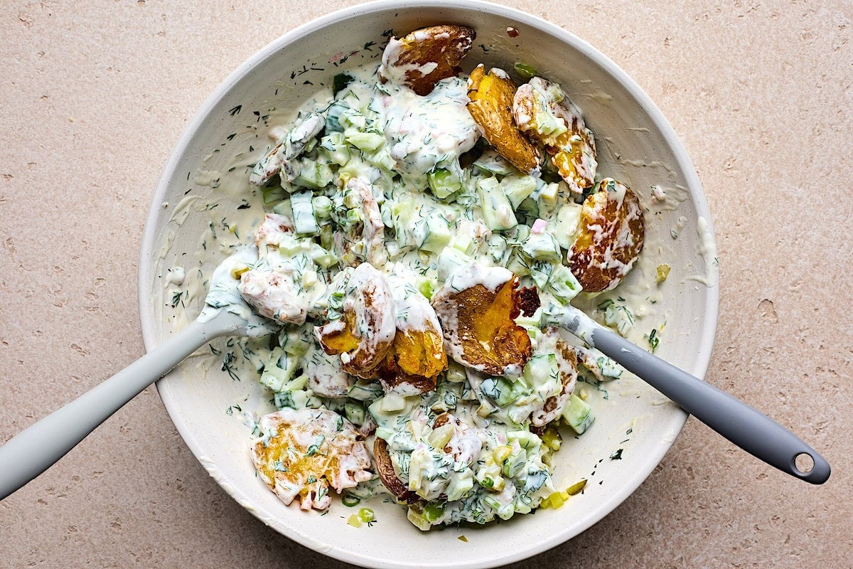 smashed potato salad being tossed in creamy herb dressing in bowl.
