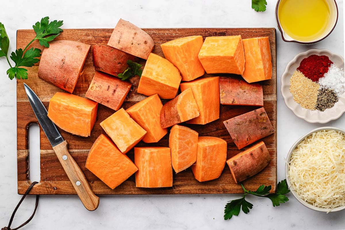 sweet potatoes cut into large chunks on wood cutting board with knife. 