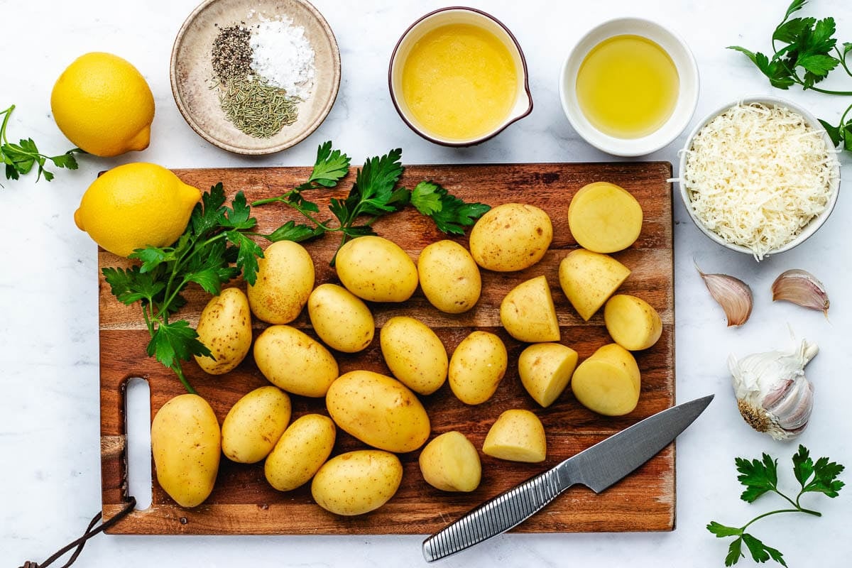 Yukon gold potatoes on cutting board with knife with lemons, parsley, garlic, spices, melted butter, oil, and Parmesan cheese.