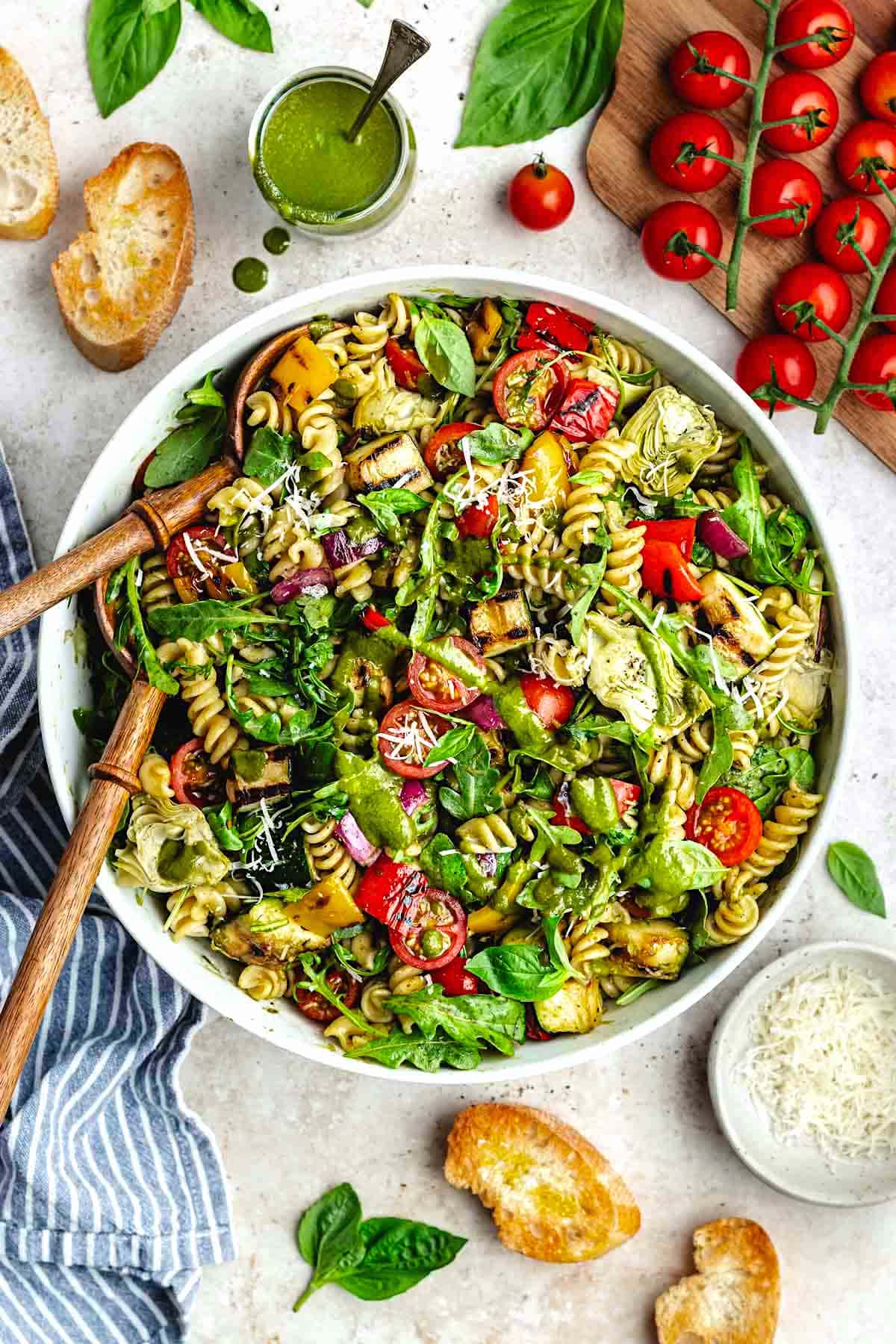 grilled vegetable pasta salad with basil vinaigrette and parmesan cheese in white serving bowl with wood salad servers.