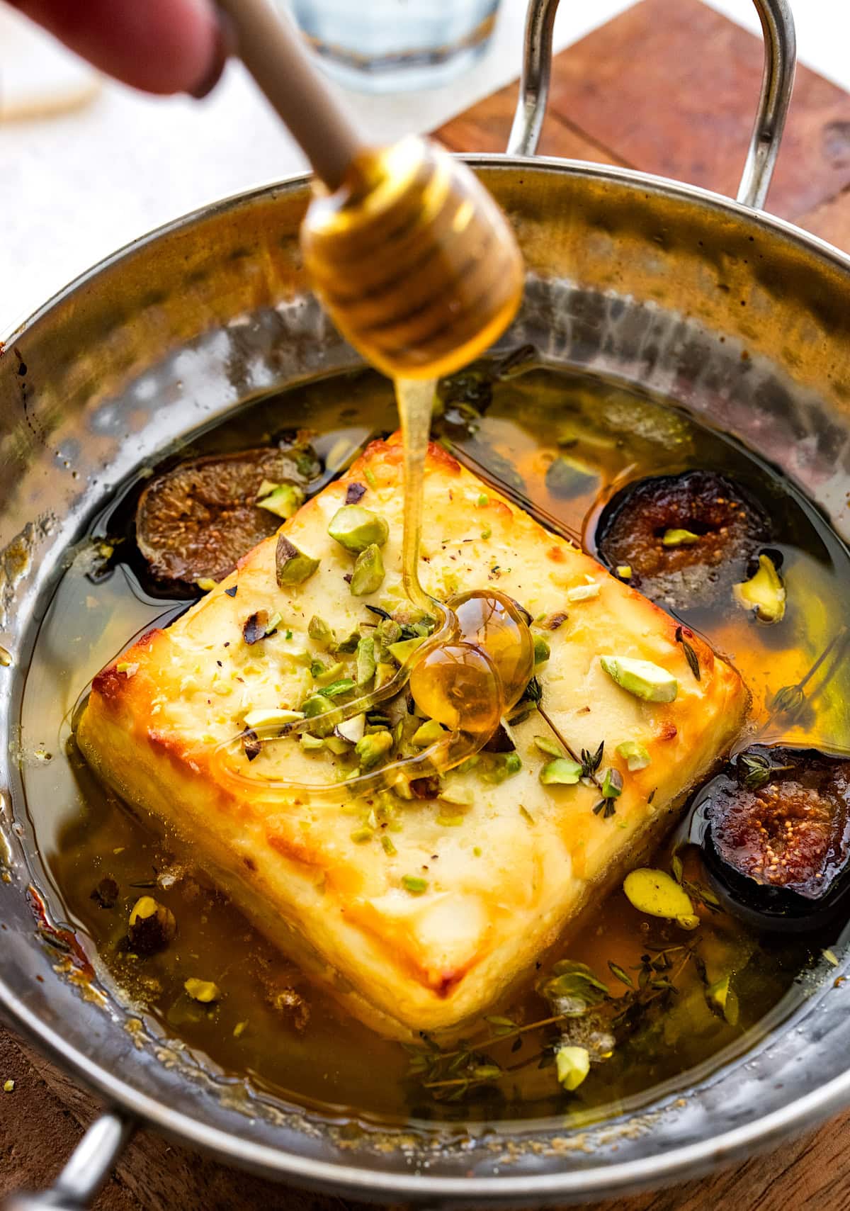 baked feta in dish with pistachios, figs, thyme, and honey being drizzled over the top.