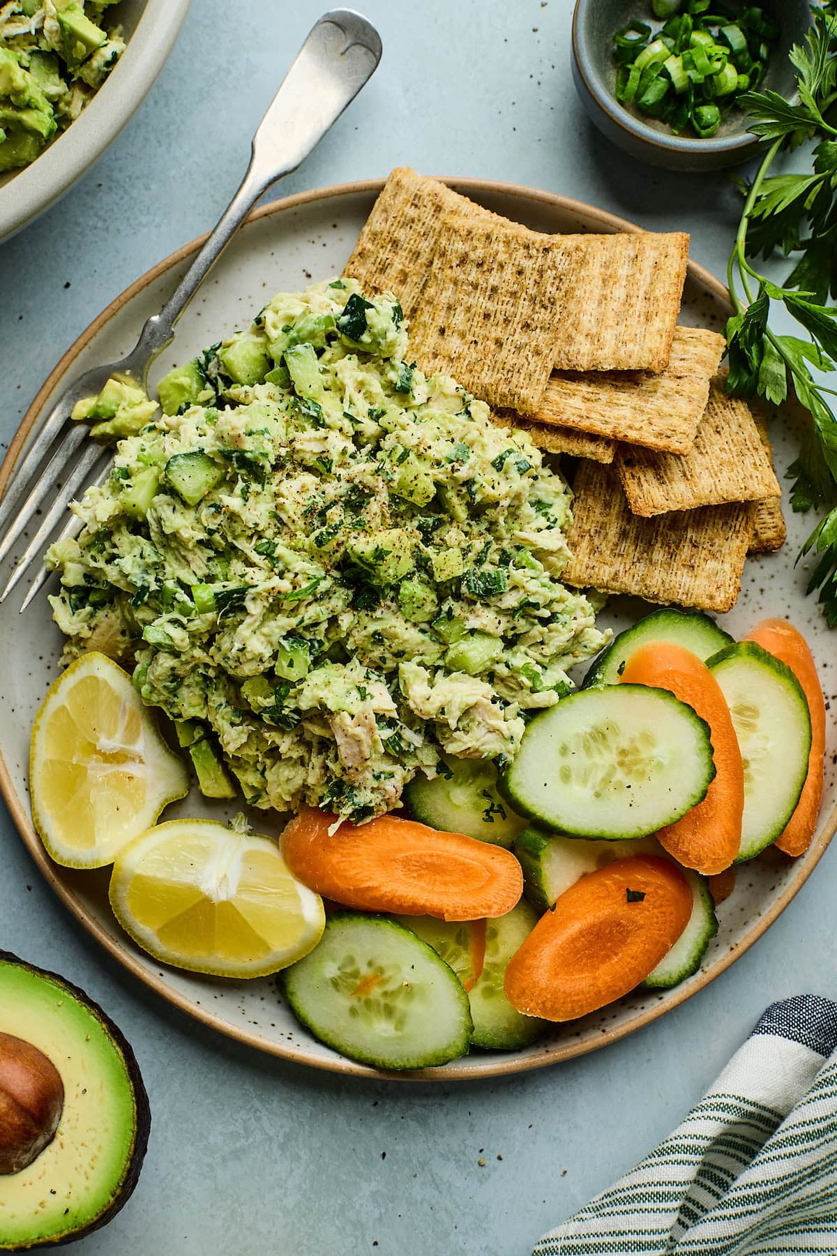 avocado chicken salad on plate with crackers, cucumbers, carrots, lemon wedges, and fork.